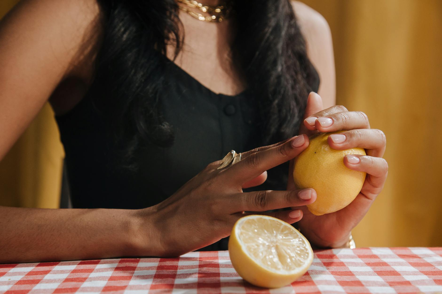 A hand with white nails holding a fresh lemon on a soft pink background surrounded by additional lemons, representing the duality of solo and shared pleasure.