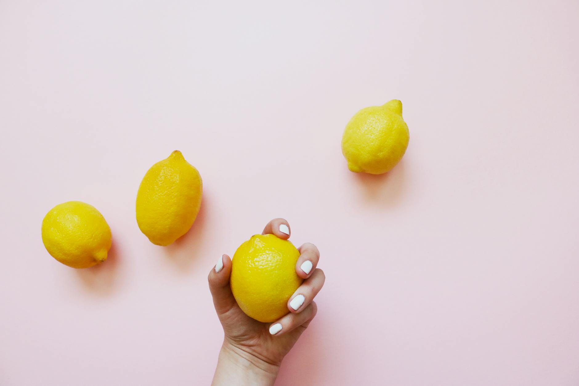 A hand with white nails holding a fresh lemon on a soft pink background, surrounded by additional lemons.