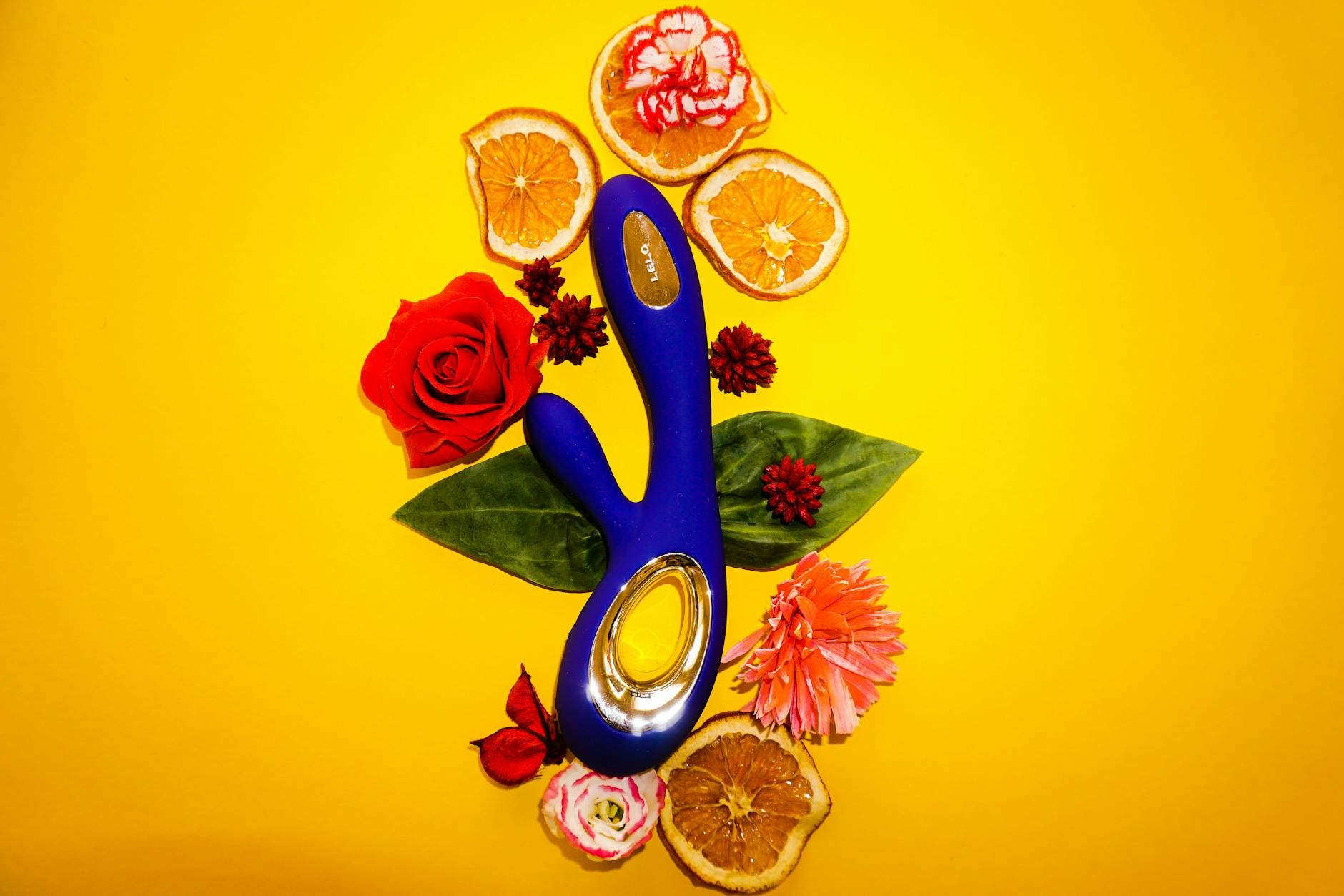 A blue vibrator surrounded by flowers and citrus on a yellow background